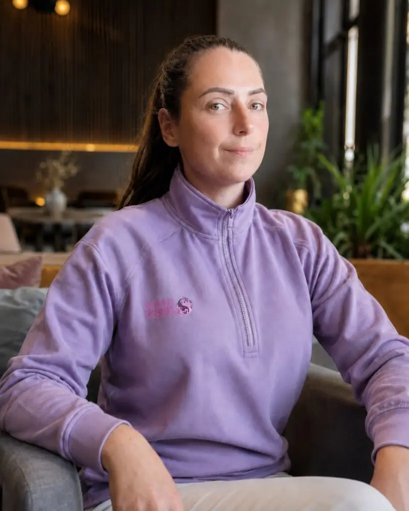 Woman wearing a lilac fleece zip-up sits on a sofa in a cafe, looking at the camera with a calm expression.